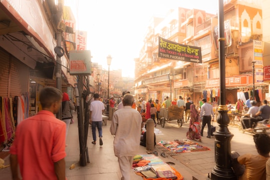 A bustling street scene featuring numerous people walking and shopping. The street is lined with various shops, displaying colorful textiles and items for sale. The architecture showcases traditional designs with ornate details. There is a cart being pushed and a street sign advertising tour packages.