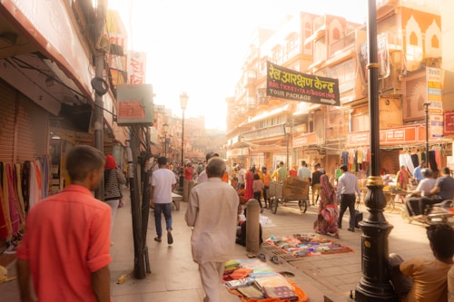 A bustling street scene featuring numerous people walking and shopping. The street is lined with various shops, displaying colorful textiles and items for sale. The architecture showcases traditional designs with ornate details. There is a cart being pushed and a street sign advertising tour packages.
