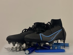 A pair of black and blue soccer cleats with metal studs, placed on top of a blister pack of capsules and several blue vape pens. The cleats have a sleek design with blue accents and appear to be of professional quality.