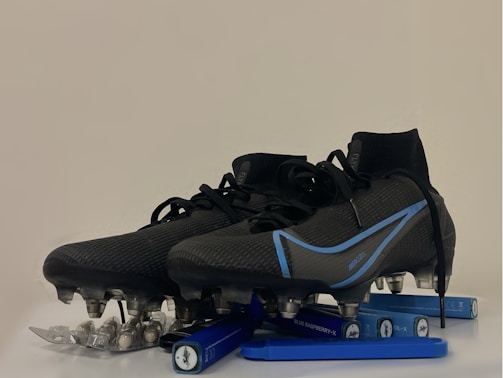A pair of black and blue soccer cleats with metal studs, placed on top of a blister pack of capsules and several blue vape pens. The cleats have a sleek design with blue accents and appear to be of professional quality.