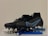 A pair of black and blue soccer cleats with metal studs, placed on top of a blister pack of capsules and several blue vape pens. The cleats have a sleek design with blue accents and appear to be of professional quality.