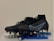 A pair of black and blue soccer cleats with metal studs, placed on top of a blister pack of capsules and several blue vape pens. The cleats have a sleek design with blue accents and appear to be of professional quality.
