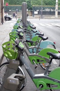 A row of rental bicycles neatly lined up in a city bike-sharing station, with green and grey color schemes. The background features a street with a pedestrian crossing and green trees behind a black iron fence. There are people walking and cars on the street, indicating an urban environment.