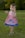 A young child wearing a red and white checkered dress with a blue trim is standing on a grassy field. The dress features a cartoon whale design. The child has short brown hair and is barefoot, holding their hands up and smiling. The background consists of blurred green trees, creating a natural setting.