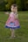 A young child wearing a red and white checkered dress with a blue trim is standing on a grassy field. The dress features a cartoon whale design. The child has short brown hair and is barefoot, holding their hands up and smiling. The background consists of blurred green trees, creating a natural setting.