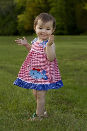 A young child wearing a red and white checkered dress with a blue trim is standing on a grassy field. The dress features a cartoon whale design. The child has short brown hair and is barefoot, holding their hands up and smiling. The background consists of blurred green trees, creating a natural setting.