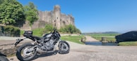 A modern motorcycle is parked on a gravel path near an old, picturesque stone castle surrounded by lush greenery. The sky is clear and blue, and a small stream flows under a stone bridge leading towards the castle. The landscape includes rolling hills in the distance.