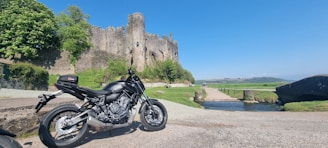 A modern motorcycle is parked on a gravel path near an old, picturesque stone castle surrounded by lush greenery. The sky is clear and blue, and a small stream flows under a stone bridge leading towards the castle. The landscape includes rolling hills in the distance.