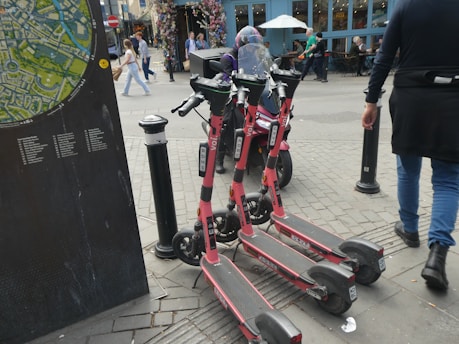 Several electric scooters are parked on a city sidewalk near a public map display. Pedestrians walk by, and some people are seated at tables outside a nearby cafe. The scene is urban and bustling with activity.