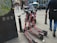 Several electric scooters are parked on a city sidewalk near a public map display. Pedestrians walk by, and some people are seated at tables outside a nearby cafe. The scene is urban and bustling with activity.