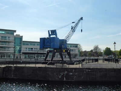 A large, blue industrial crane is positioned on a dockside with modern white buildings in the background. The crane appears to be stationary, and there is a body of water in the foreground. The scene is bright and clear with a blue sky overhead.