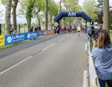 A group of people gathering at a starting line under an inflatable archway for what appears to be a running event. The arch bears the name 'AIN' and is surrounded by tall trees, suggesting a park or tree-lined street setting. Spectators are seen on the sidelines, some capturing the event using cameras or phones.