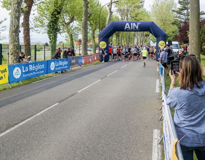 A group of people gathering at a starting line under an inflatable archway for what appears to be a running event. The arch bears the name 'AIN' and is surrounded by tall trees, suggesting a park or tree-lined street setting. Spectators are seen on the sidelines, some capturing the event using cameras or phones.