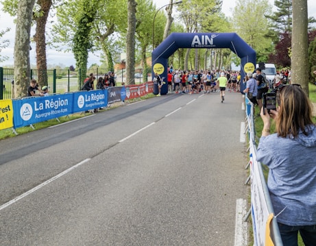 A group of people gathering at a starting line under an inflatable archway for what appears to be a running event. The arch bears the name 'AIN' and is surrounded by tall trees, suggesting a park or tree-lined street setting. Spectators are seen on the sidelines, some capturing the event using cameras or phones.