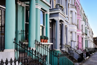 A row of colorful terraced houses lines a street. Each house is painted in different pastel shades, including teal, pink, and light purple. The houses feature classical architectural elements such as columns and ornate iron railings. Potted plants can be seen on some of the window sills, and the pavement in front is neatly arranged with large grey stones.