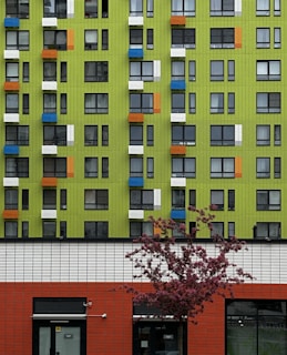 A modern apartment building with a vivid green facade is accented by scattered blue, orange, and white window panels. In the foreground, a red brick section of the building contrasts with the green upper floors, and a blooming pink tree stands out against the clean geometric lines of the architecture.