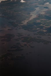 An aerial view of a landscape featuring a network of waterways and patches of land. The image shows various small islands surrounded by a large body of water. Scattered clouds cast shadows on the surface below, while different shades of green and brown indicate forested and open land areas.