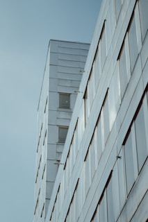 A modern building with a sleek, minimalistic design featuring reflective glass windows and smooth, clean lines. The structure appears to rise into the sky, with a clear, light blue background enhancing its contemporary aesthetic.