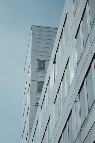 A modern building with a sleek, minimalistic design featuring reflective glass windows and smooth, clean lines. The structure appears to rise into the sky, with a clear, light blue background enhancing its contemporary aesthetic.