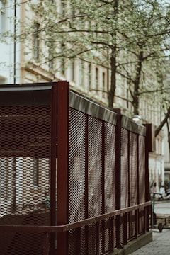 A metal mesh fence with a reddish-brown frame is set alongside a city street. Behind the fence, a building with classic architecture can be seen, along with a tree with sparse leaves. The scene suggests an urban setting with paved sidewalks.