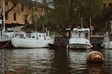 Several boats are moored along a tree-lined embankment, with an old building in the background. The water's surface is calm, reflecting the muted colors of the scene. The environment suggests a serene and possibly historic place, with one boat displaying a flag and a life ring hanging on a nearby pole.