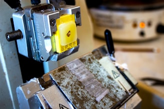 A close-up view of a microtome, featuring a yellow sample holder attached to the machine. The setup includes metallic parts and a partially visible blade segment. The background shows blurred laboratory equipment, including a container with controls and a pencil.