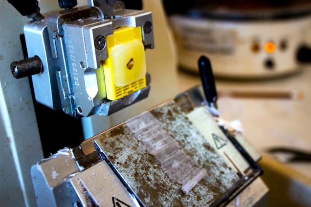 A close-up view of a microtome, featuring a yellow sample holder attached to the machine. The setup includes metallic parts and a partially visible blade segment. The background shows blurred laboratory equipment, including a container with controls and a pencil.