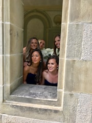 Five women dressed in navy blue outfits are posing together within a stone-framed window. They are smiling and appear to be part of a celebratory occasion, likely a wedding. Behind them, there are ornate wall decorations and some white flowers are visible.
