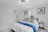 A bedroom featuring a neatly made bed with a white comforter and blue throw blanket. Geometric-patterned pillows complement the modern art on the walls. A small closet with a hanging rack and ladder is to the side. Two small bedside tables with potted plants add a touch of greenery.