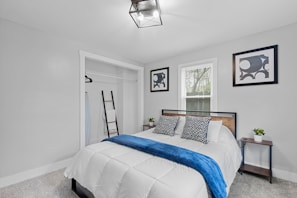 A bedroom featuring a neatly made bed with a white comforter and blue throw blanket. Geometric-patterned pillows complement the modern art on the walls. A small closet with a hanging rack and ladder is to the side. Two small bedside tables with potted plants add a touch of greenery.
