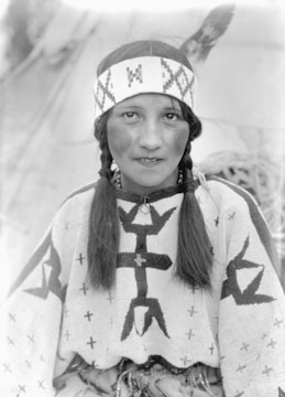 A young person with braided hair is wearing a headband with geometric patterns and an intricately decorated garment featuring crosses and fish designs. There is a feather attached to the headband. The background appears to be a blurred outdoor setting.