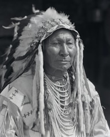 A person wearing a traditional headdress adorned with feathers and beads. The individual appears to be dressed in ceremonial attire, including a beaded necklace and a garment with decorative patterns.
