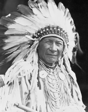 An individual wearing a traditional headdress adorned with large feathers and ornate patterns. The person is dressed in elaborate tribal clothing, featuring multiple beaded necklaces and decorations made from natural materials. The image is in black and white, enhancing the focus on the textures and intricate details of the attire.