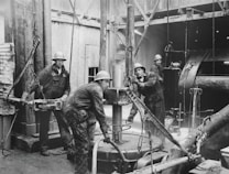 Four workers are engaged in an industrial setting, possibly an oil rig or construction site. They are wearing protective clothing and helmets, surrounded by heavy machinery and equipment. The environment suggests manual labor and teamwork.