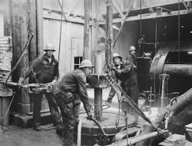 Four workers are engaged in an industrial setting, possibly an oil rig or construction site. They are wearing protective clothing and helmets, surrounded by heavy machinery and equipment. The environment suggests manual labor and teamwork.
