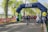 A group of people, including participants in running gear and costumed characters, gather at a starting line under a large inflatable arch. The arch is blue with promotional writing, while the surrounding area is green with trees and a clear sky. Security barriers flank the path, and there are banners advertising a marathon event.