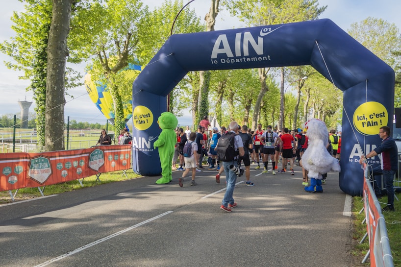 A group of people, including participants in running gear and costumed characters, gather at a starting line under a large inflatable arch. The arch is blue with promotional writing, while the surrounding area is green with trees and a clear sky. Security barriers flank the path, and there are banners advertising a marathon event.