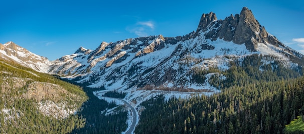 A breathtaking mountain landscape with snow-covered peaks and rugged formations. A winding road cuts through dense forests, leading the eye towards the towering mountains under a clear blue sky.