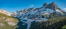 A breathtaking mountain landscape with snow-covered peaks and rugged formations. A winding road cuts through dense forests, leading the eye towards the towering mountains under a clear blue sky.