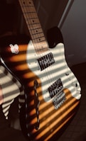 A close-up of an electric guitar with a sunburst finish. The guitar features a classic single cutaway shape with visible pickups and control knobs. Sunlight creates striped shadows across the guitar body, enhancing its wooden texture.