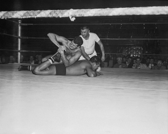 In a boxing ring, two wrestlers are engaged in a match while a referee watches closely. One wrestler has the other pinned down on the mat, demonstrating strength and technique. The audience in the background observes the match from the stands with interest.