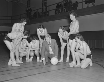 A group of female basketball players in uniforms gathers around a coach who is crouching near a basketball. They appear to be strategizing or planning, with the coach pointing at the court. The background includes a balcony with people observing the scene.