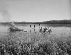 A group of people sitting on chairs in a semi-circle on an ice-covered lake, with a pile of branches burning nearby. Tall grass lines the foreground, and a forest stretches along the horizon under a clear sky. Two individuals are standing separately on the ice, one of them holding a hockey stick.