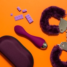 An arrangement of objects on an orange background includes purple handcuffs with a furry texture, a purple ergonomic device, a purple pouch, and several purple plastic bricks resembling toy blocks.
