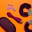 An arrangement of objects on an orange background includes purple handcuffs with a furry texture, a purple ergonomic device, a purple pouch, and several purple plastic bricks resembling toy blocks.
