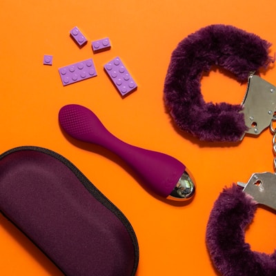 An arrangement of objects on an orange background includes purple handcuffs with a furry texture, a purple ergonomic device, a purple pouch, and several purple plastic bricks resembling toy blocks.