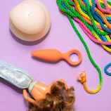 A pastel purple background features a large cream-colored candle, a multicolored bundle of rope with green, pink, yellow, and blue hues, a small orange object with a ring handle, and a plastic doll with curly brown hair and iridescent clothing.