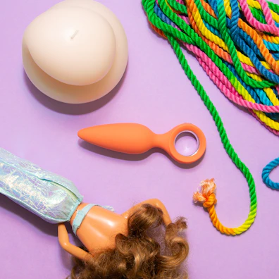 A pastel purple background features a large cream-colored candle, a multicolored bundle of rope with green, pink, yellow, and blue hues, a small orange object with a ring handle, and a plastic doll with curly brown hair and iridescent clothing.