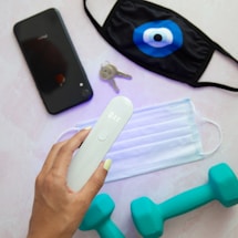 A hand holding a white electronic device with an LED display. Beside it are a black smartphone, two keys, a black face mask with a blue eye design, a surgical mask, and two teal dumbbells on a light pink surface.