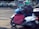 A person wearing a colorful helmet and dark jacket is seated on a white scooter parked at a crosswalk. The scooter is equipped with a large red cover, and there is a blue plastic bag on the footrest. Vehicles are visible in the background, along with a graffiti-covered sign.